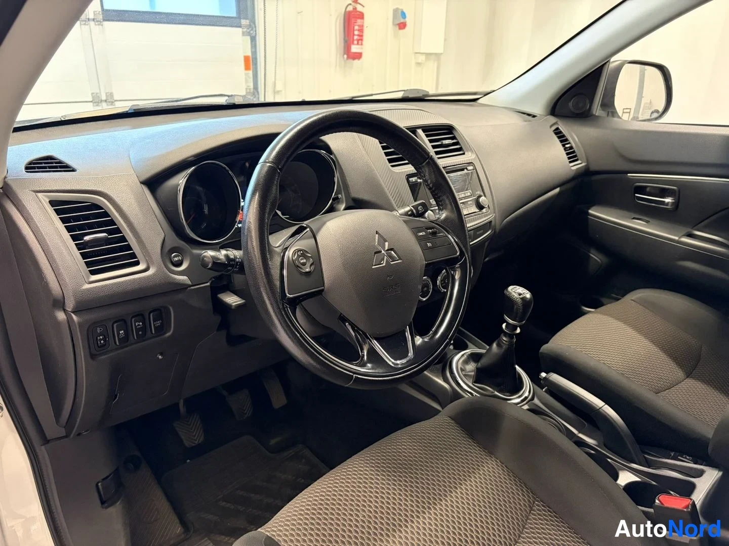 Yksityiskohta: Auton ohjaamo kuljettajan puolelta kuvattuna: Mitsubishi-merkillinen ohjauspyörä, analogiset mittarit, käytetty manuaalivaihteisto ja kankaat penkit, vasemmalla