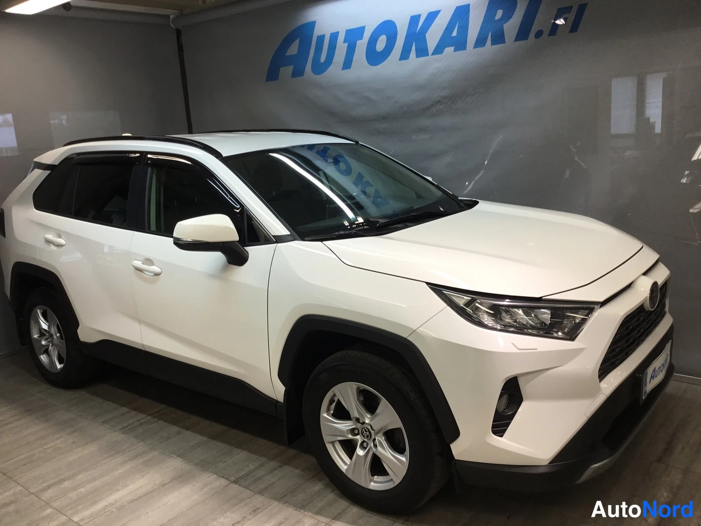 Etuviisto, nokka oikealle: Valkoisen Toyota RAV4 -maasturin etukulmanäkymä sisätiloissa; etusäleikkö ja ajovalot näkyvät oikealla, kylki vasemmalla.