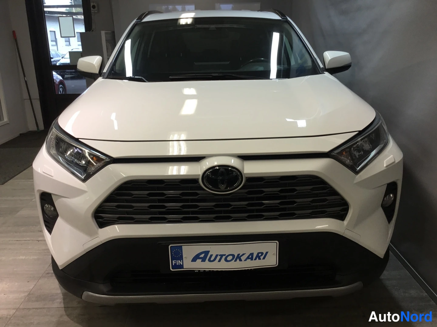 Edestä: Toyotan etupää edestä: valkoinen koripaneeli, Toyota-merkki grillessä ja Autokari-kilpi rekisteripaikalla.