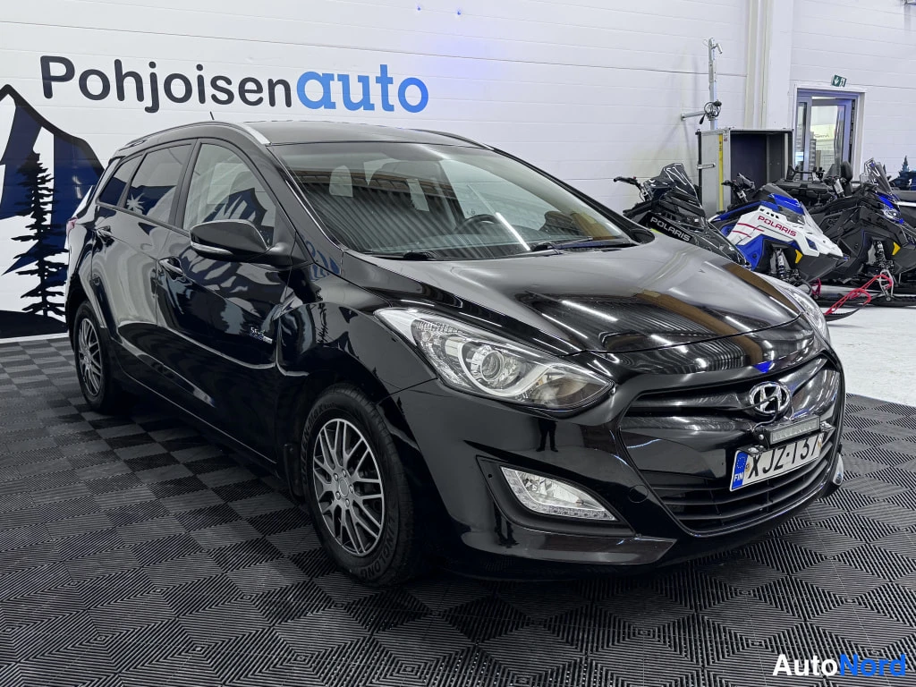 Etuviisto, nokka oikealle: Mustan Hyundai i30 -auton oikeanpuoleinen etukulmanäkymä, rekisterikilpi, taustalla lumikelkkoja ja Pohjoisauto-mainostaulu.