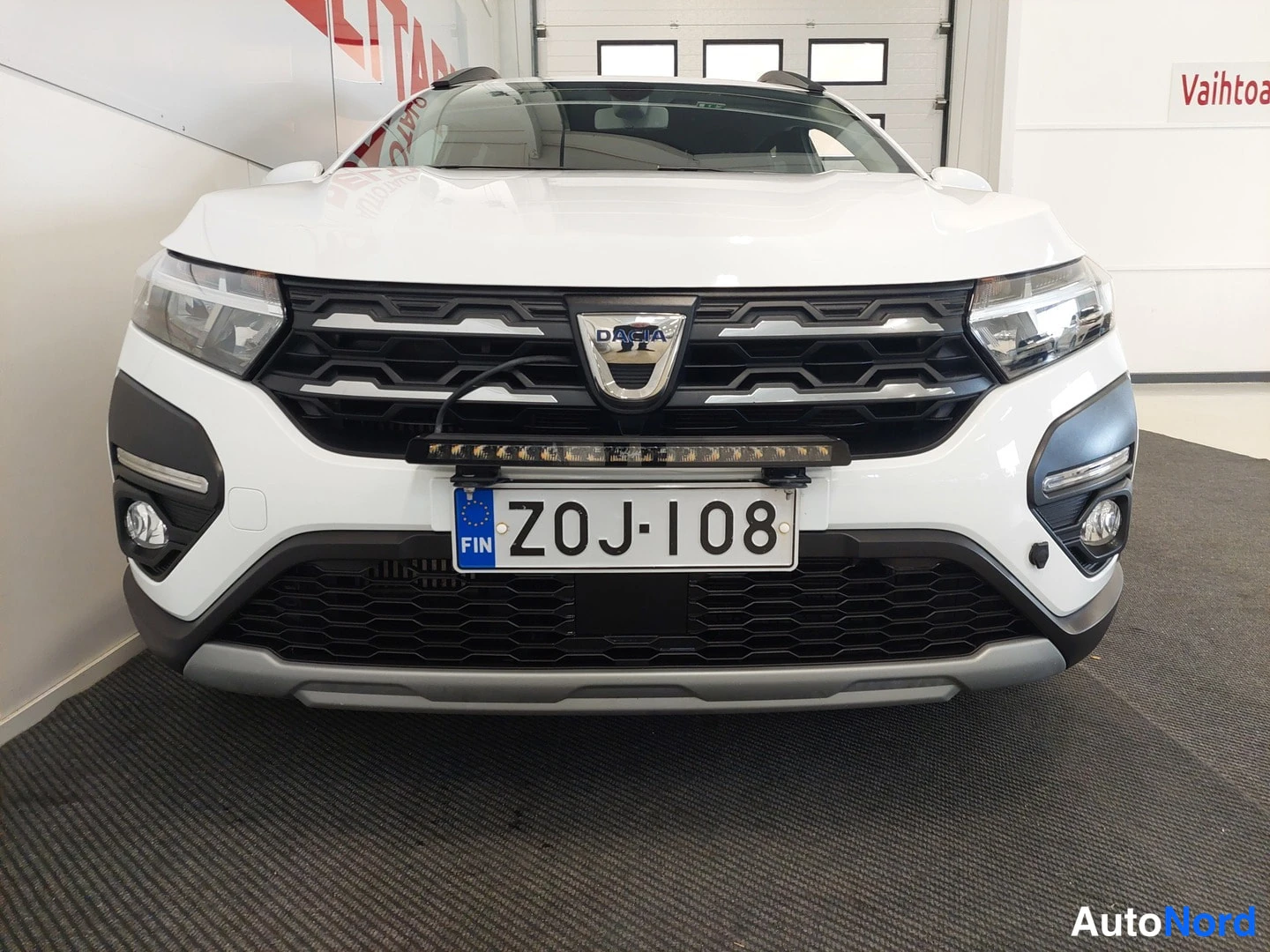 Edestä: Auton etupää: Dacia-merkki keulassa, suorakulmainen LED-tyyppinen lisävalo etupuskurin yläpuolella, rekisteritunnus.