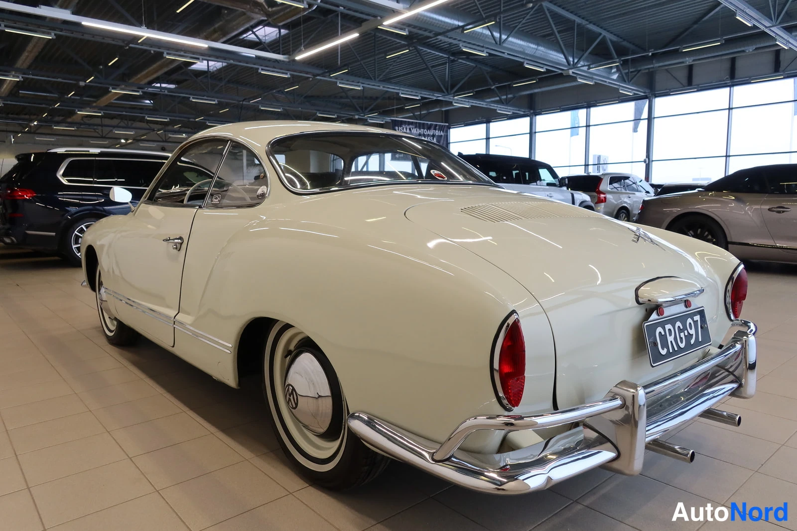 Takaviisto: Kermanvalkoinen Volkswagen Karmann-Ghia -coupe takaviistosta rättikäshallissa; rekisterikilpi, kromattu puskuri ja punaiset takavalot näkyvissä.