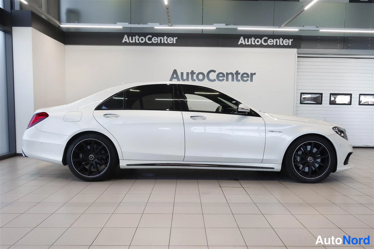 Sivulta: Valkoinen Mercedes-Benz S 63 AMG -sedan sivuprofiili sisätiloissa, mustat kevytmetallivanteet ja kiiltävä katto.