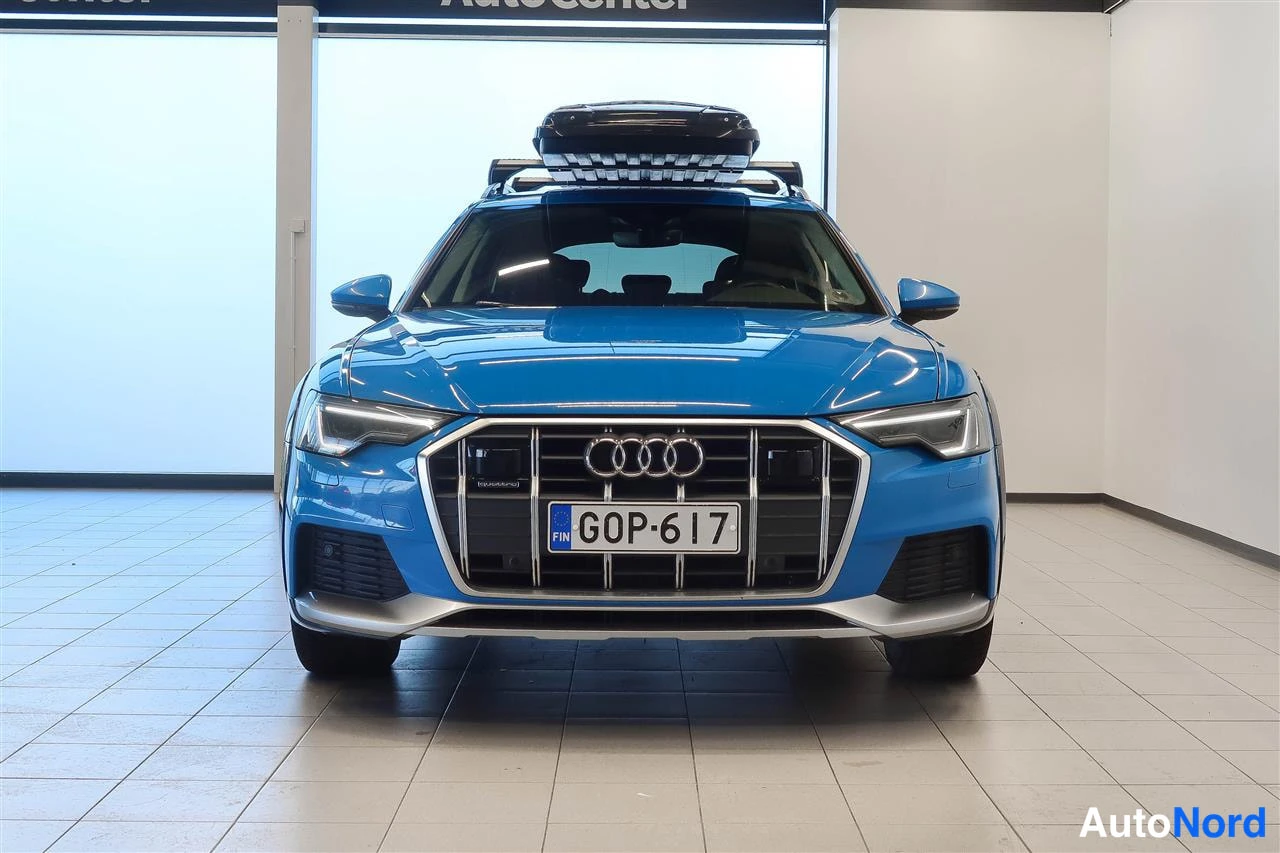 Edestä: Sininen auto suoraan edestä päin, Audi-merkki kiiltoniskassa, rekisterikilpi, musta kattolaatikko katolla.