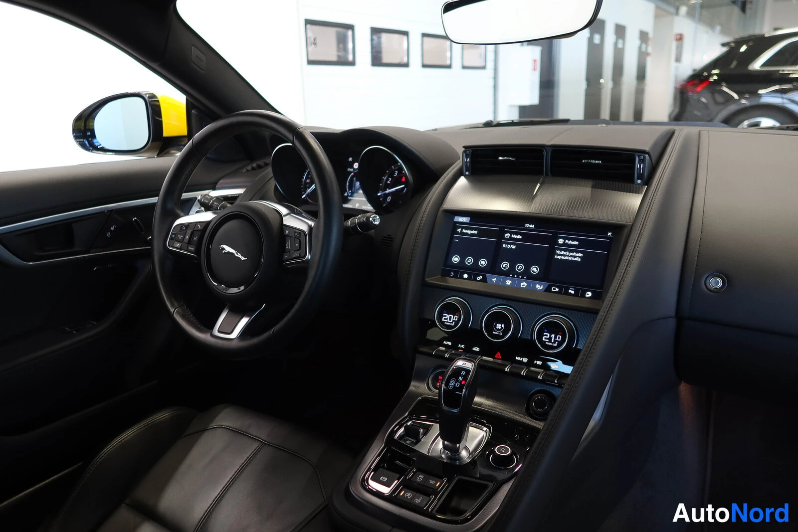 Muu: Ohjaamo kuvattuna keskikäytävältä: Jaguar-ohjauspyörä, mittarit, kosketusnäyttö ja automaattivaihteen valitsin.