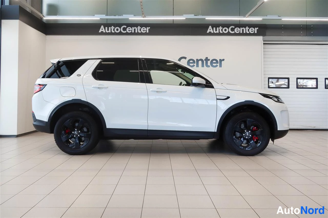 Sivulta: Land Rover Discovery Sport -maastauton sivukulma, valkoinen kori, mustat vanteet, Autokeskuksen sisätiloissa.