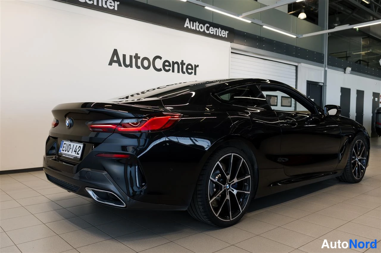 Takaviisto: Musta coupé näkyy takaa sivusuunnassa AutoCenter-tilassa; rekisterikilpi, takavalot ja oikean puolen vanteet näkyvät.