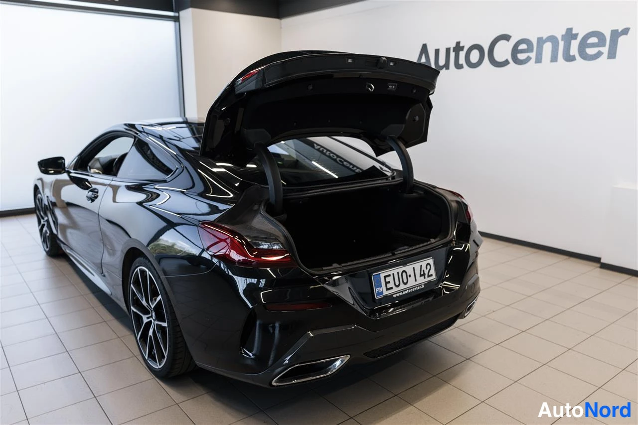 Takaviisto: Musta coupé avoimella tavaratilan kannella AutoCenter-hallissa; tyhjä tavaratila ja rekisterikilpi näkyvät.