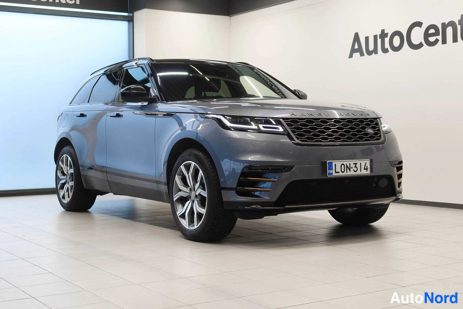Etuviisto, nokka oikealle: Hopeanharmaa Range Rover Velar -maasturi esillä autonäyttelytilassa, etukulma vasemmalle.