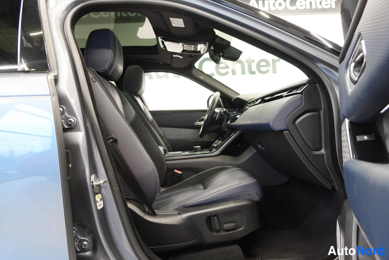 Muu: Etumatkustustila avatusta oikeanpuoleisesta ovesta kuvattuna; tummansiniset istuimet, ohjauspyörä ja keskikonsoli näkyvät taustalla AutoCenter-kyltti.
