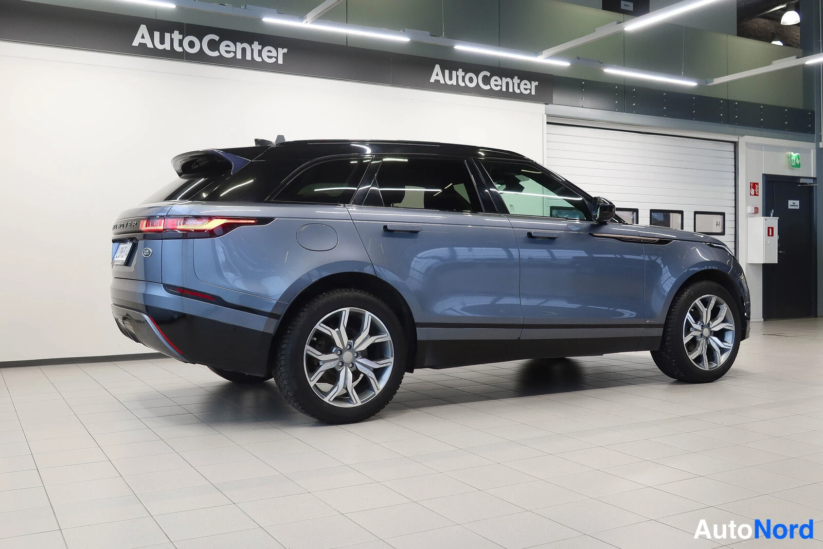 Takaviisto: Sinisenharmaan Range Rover Velar -maasturin takaosa ja vasen kylki sisätiloissa; näkyvissä takavalot, takapaneeli, musta katto ja suuret kevytmetallivanteet.