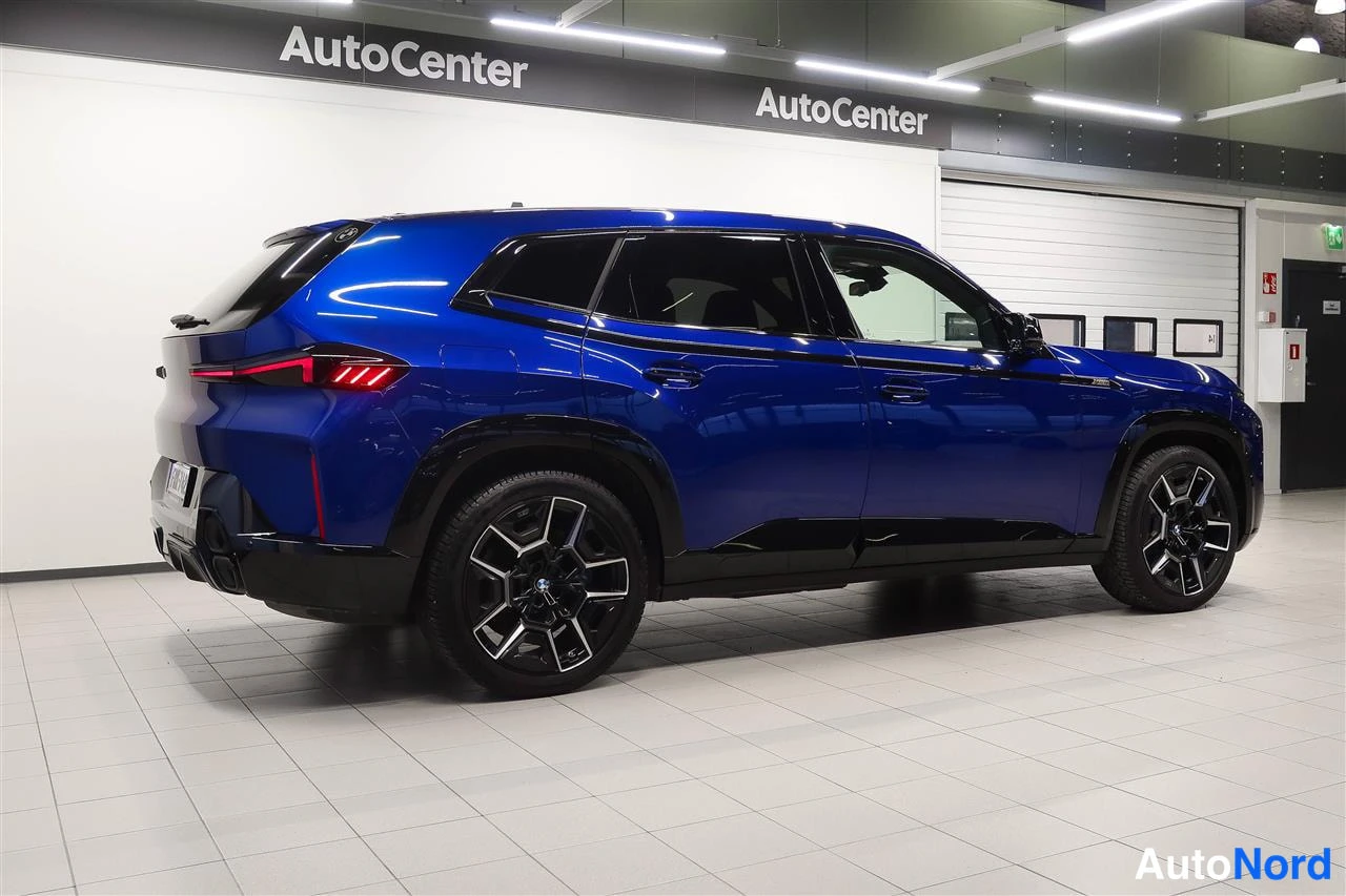 Takaviisto: Sininen BMW XM -maasturi takaviistosta autopainin sisätiloissa, takavalot ja kylki näkyvissä, mustat vanteet.