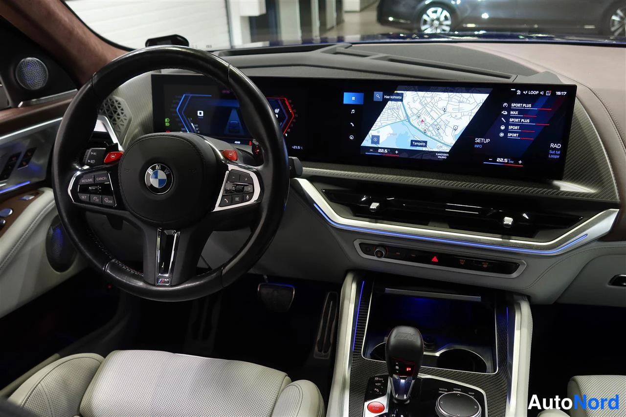 Muu: BMW XM:n ohjaamo: M-ohjauspyörä, leveä näyttöpaneeli karttanäkymällä ja ajotilan valinnat (SPORT PLUS yms.), hiilikuitukoristeet.