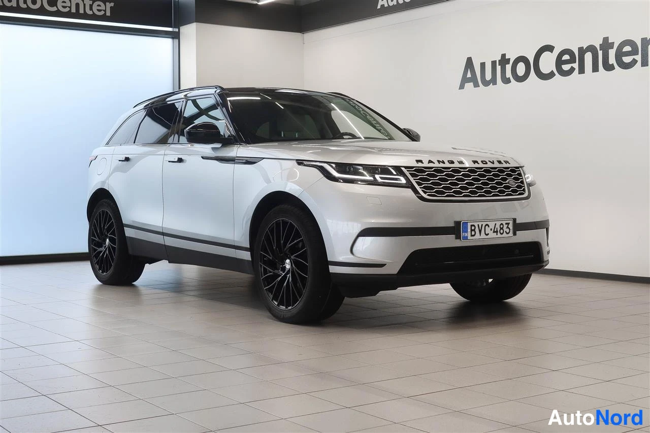 Etuviisto, nokka oikealle: Hopeanvärinen Range Rover Velar etukulmasta nähtynä AutoCenter-esittelytilassa, mustat vanteet ja -rekisterikilpi.