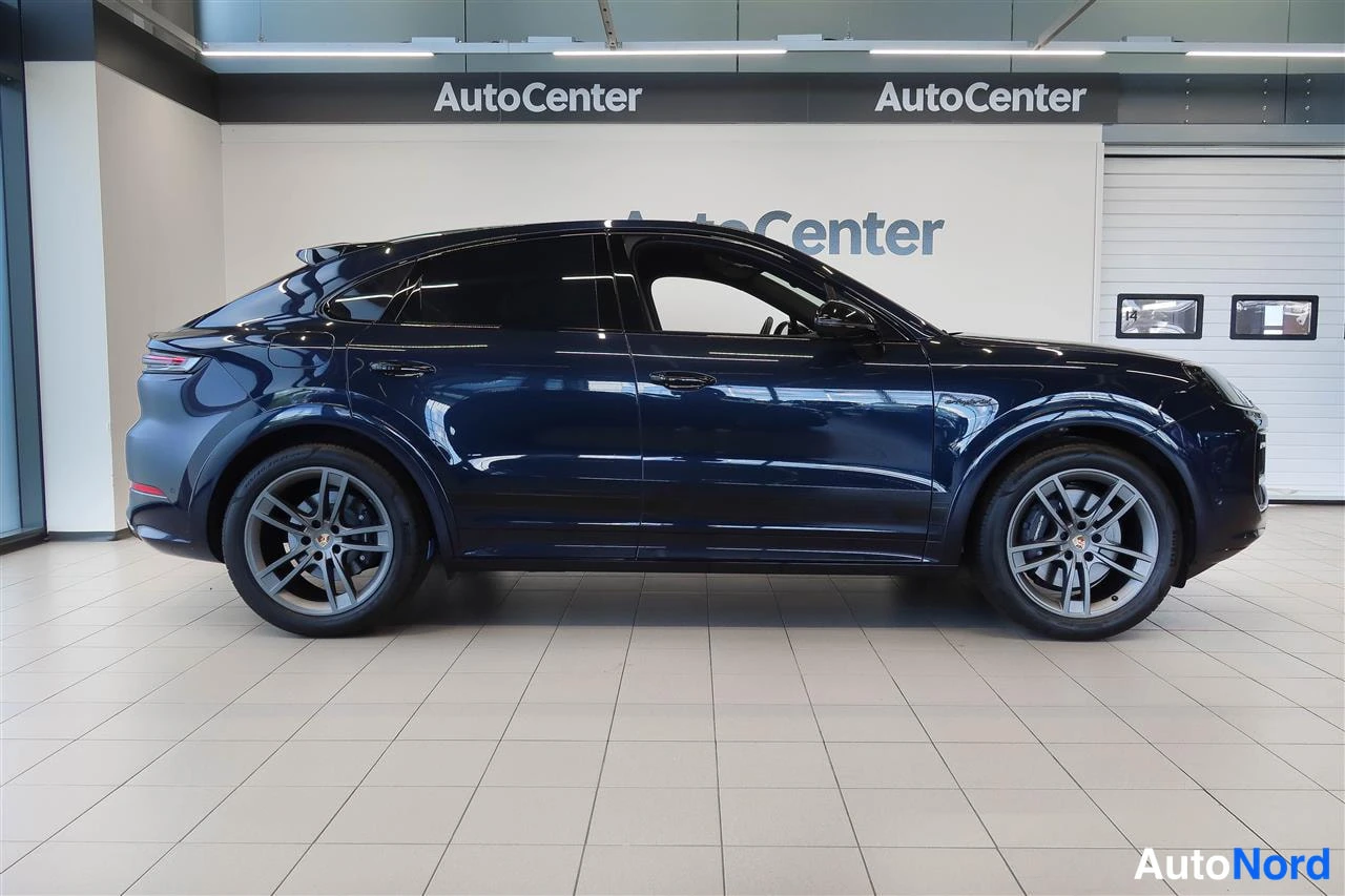 Etuviisto, nokka oikealle: Tummansininen Porsche-korinen auto sivulta kuvattuna; molemmat kyljet, pyörät ja takavalojen reuna näkyvät, taustalla AutoCenter-kyltit ja valkopaneeli.