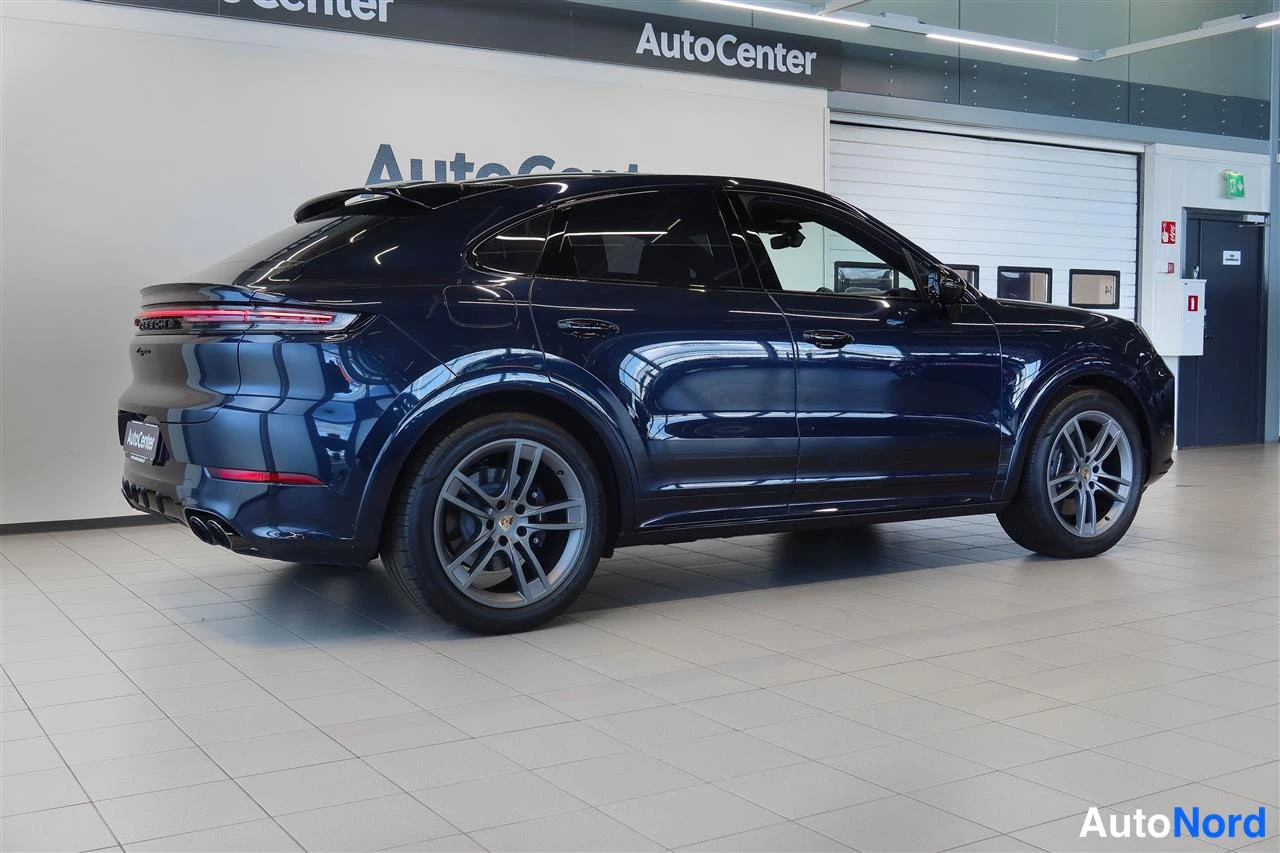 Takaviisto: Porsche Cayennen takakulma hallissa: tummansininen runko, kattosiipi ja takavalot näkyvät.