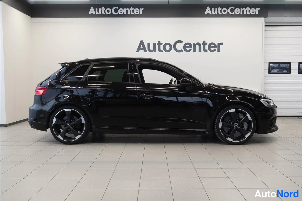 Etuviisto, nokka oikealle: Mustan Audi RS 3 Sportbackin sivukulmakuva näyttää kevytmetallivanteet, kattokiskot ja auton vasemman kyljen sisätiloissa.