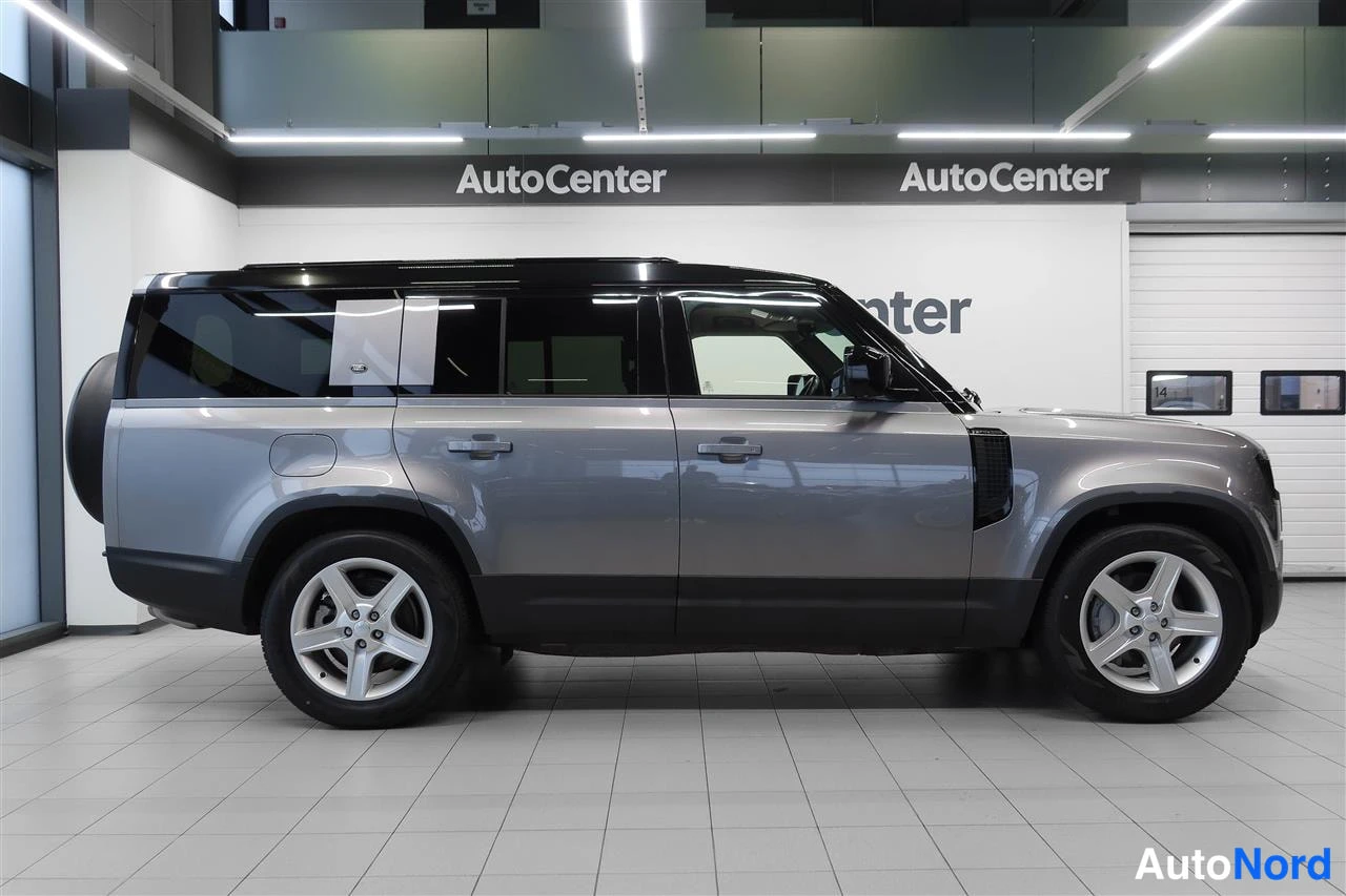 Sivulta: Land Rover Defenderin sivuprofiili hallissa, tumma katto ja harmaa koripinta, säästöpysäköintivaihtoehto taempana.
