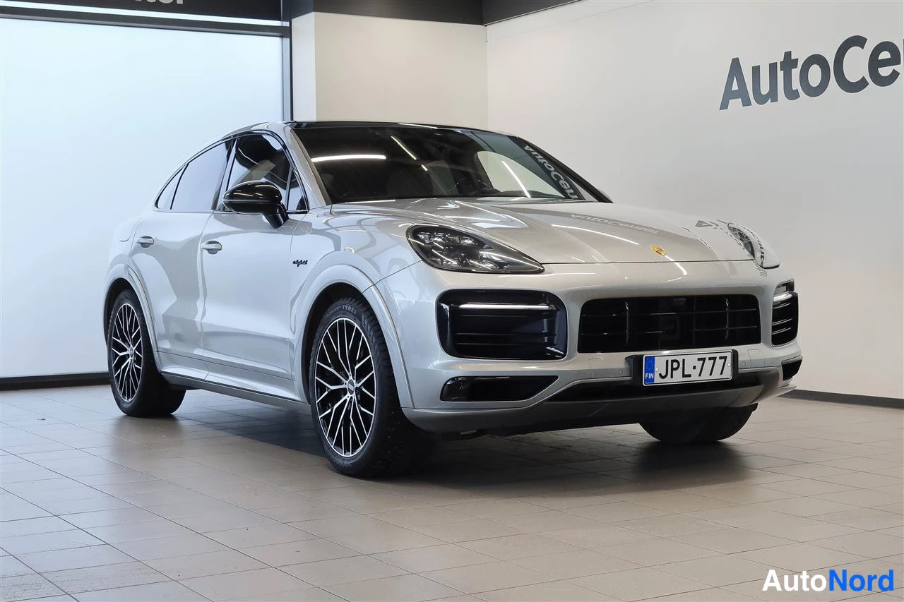 Etuviisto, nokka oikealle: Hopeinen Porsche Cayenne Coupé -tyylinen SUV esillä autosalon valkoisella lattialla, etukulmanäkyvyys oikealta, rekisteritunnus.