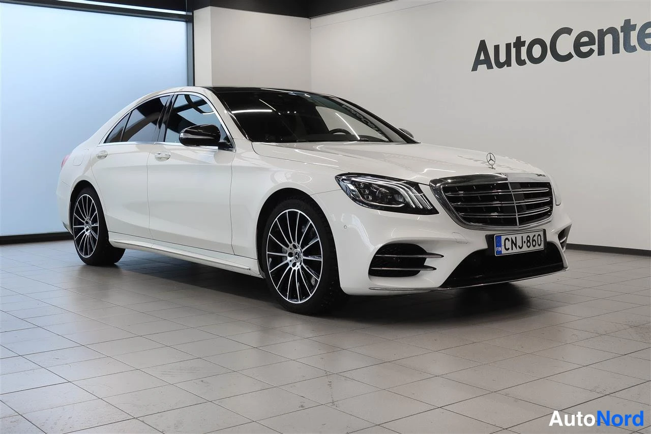 Etuviisto, nokka oikealle: Valkoinen Mercedes-Benz S-luokan sedan edestä vasemmalta kuvattuna myyntihallissa.