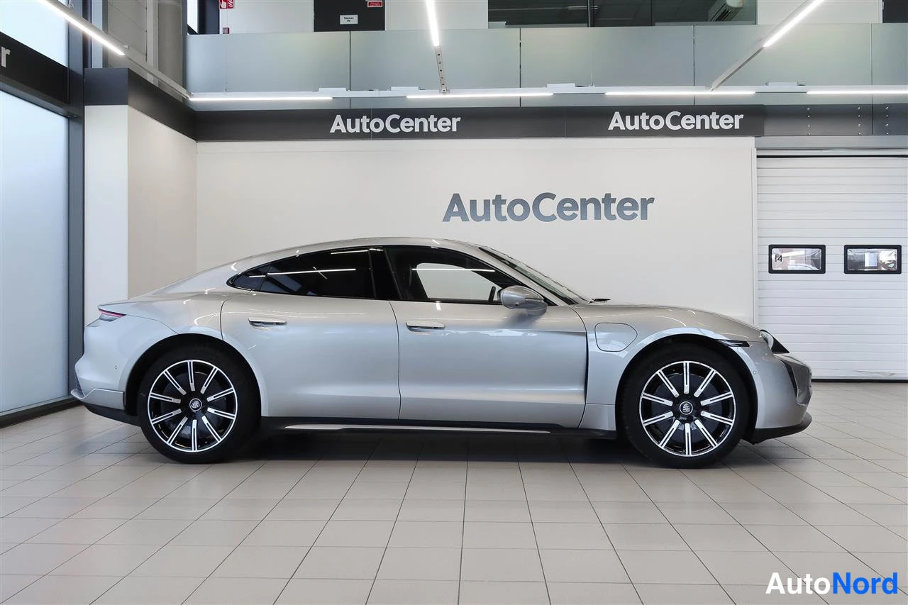 Sivulta: Porsche Taycan sivulta profiilissa, hopeanhohtoinen kori, AutoCenter-hallissa.