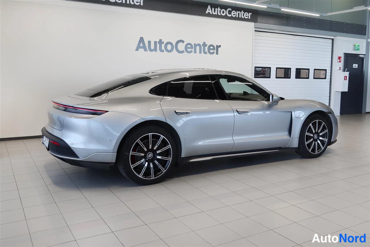Takaviisto: Hopeanvärinen Porsche Taycan -sedan takaa sivulta kuvattuna autoliikkeen showroomissa; näkyvissä takavalojen kaista, perävaunu, mustat monihaaravanteet ja punai