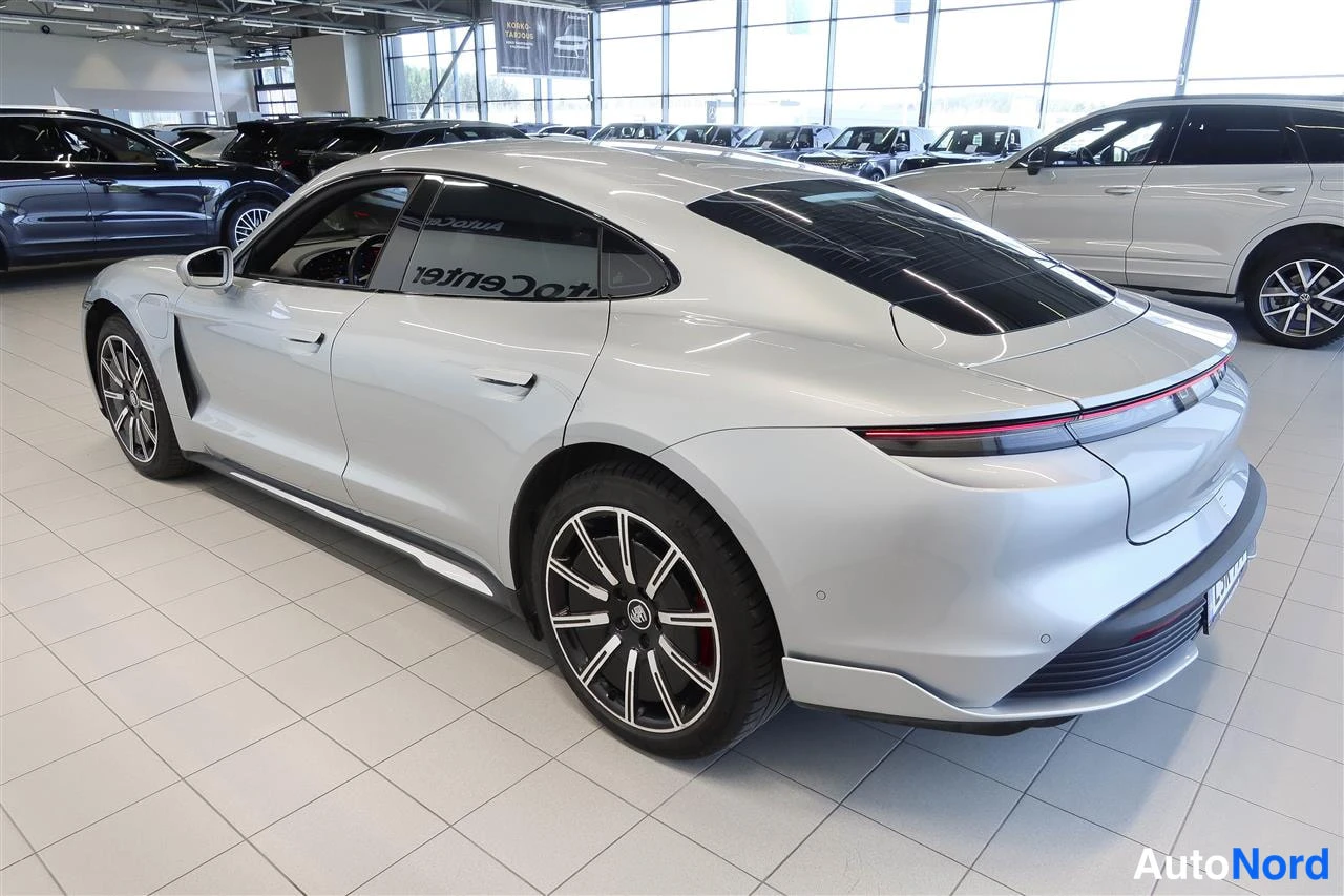 Takaviisto: Hopeanvärinen Porsche Taycan takaa kolmannesta kuvakulmasta autosalissa; peräosassa yhtenäinen takavalkokaista, tummat ikkunat ja monihaaravanteet punaisilla ja