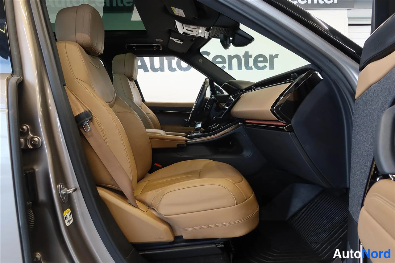 Muu: Auton sisätilan kuljettajan puolelta: beige-nahkaiset etuistuimet, mittarit ja Auto Center -tunnus taustalla.