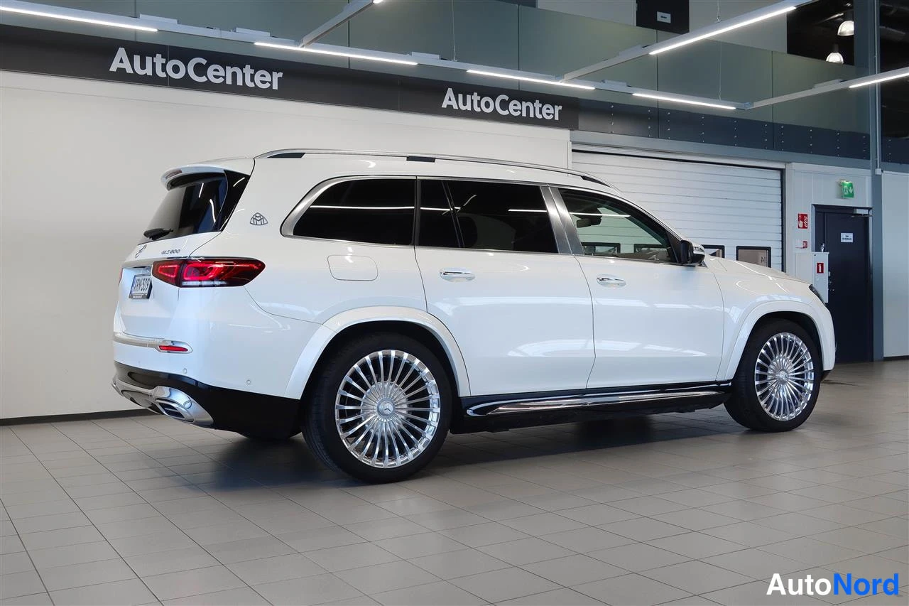 Takaviisto: Valkoinen Mercedes-Benz GLS -maasturi takaa- ja sivulta kuvattuna-autosalissa; nokka osoittaa oikealle, tummat ikkunat ja isot krominväriset vanteet.