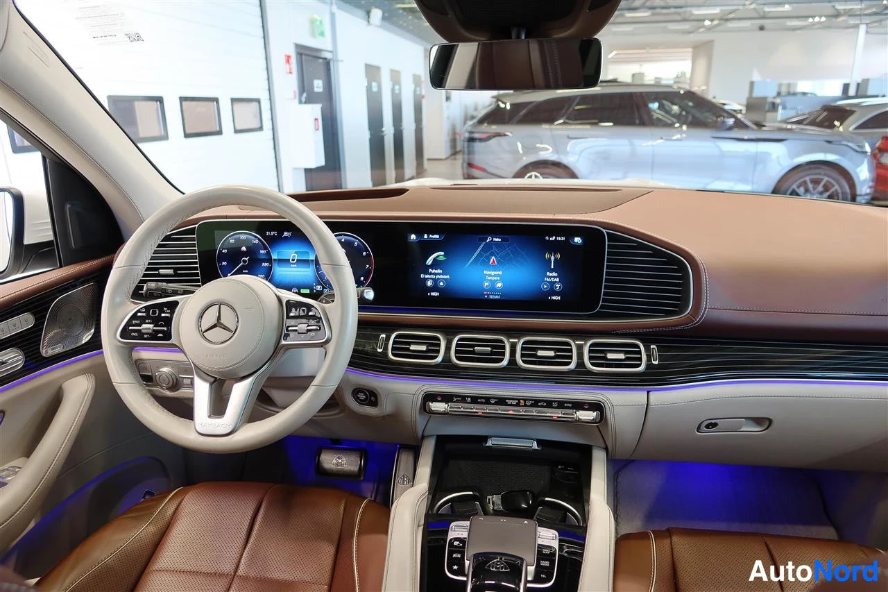 Yksityiskohta: Ohjaamosta: beige ratissa Mercedes-tähti, laaja näyttöpaneeli navigaatiokartalla, ruskeat nahkaistuimet ja sininen tunnelivalo.