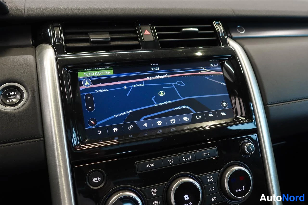 Yksityiskohta: Kohtelaulun infotainment-näyttö näyttää karttaanavigaatiota tekstillä TUTKI KARTTAA ja Paasikiventie; alla automaatti-ilmastoinnin AUTO-painikkeet ja START ENGI