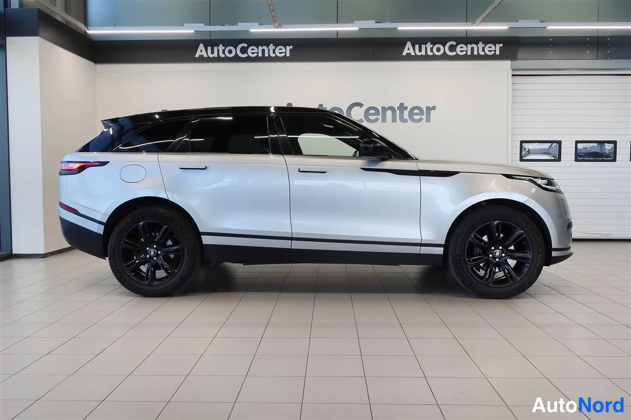 Sivulta: Sivunäkymä hopeisesta Land Rover Range Rover Velarista autohallissa, mustat vanteet ja musta kattolinja.
