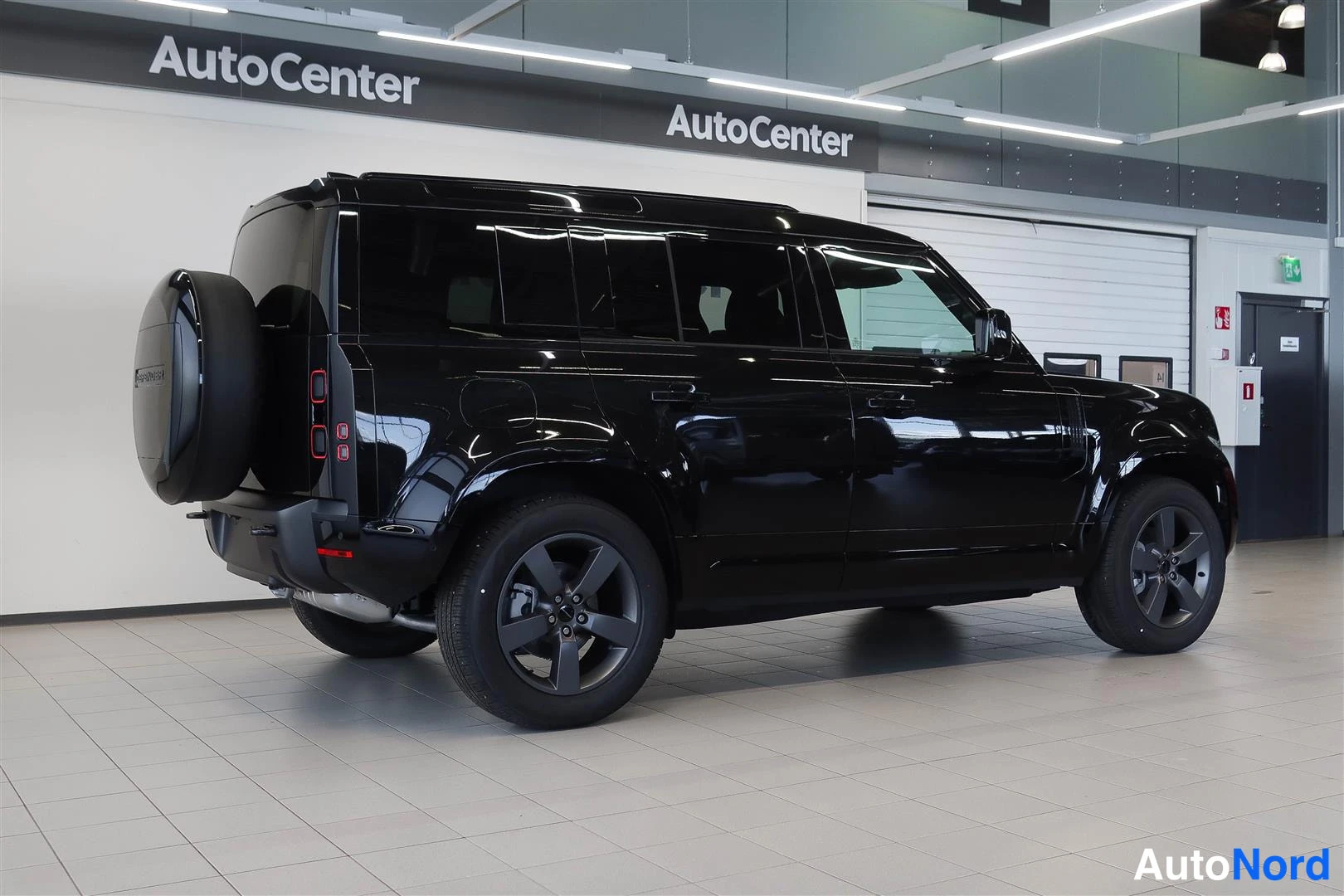 Takaviisto: Land Rover Defender 110 sivulta-takaa kuvattuna, musta kori, varavaijeri takaluukissa, tummat kevytmetallivanteet, AutoCenter-hallissa.
