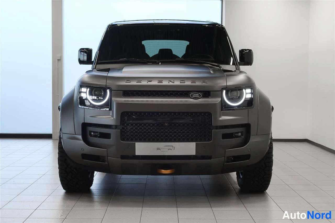 Edestä: Land Rover Defenderin suora etunäkymä tummanharmaana sisätiloissa, pyöreät ajovalot ja etusäleikkö näkyvissä.