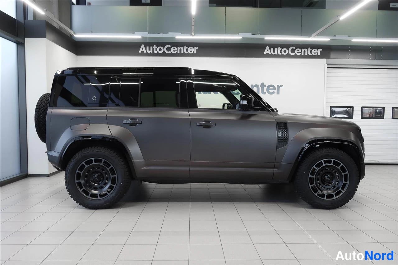 Sivulta: Land Rover Defenderin sivunäkymä tummanharmaana sisätiloissa, takarenkaiden vararahat neljällä ovella.