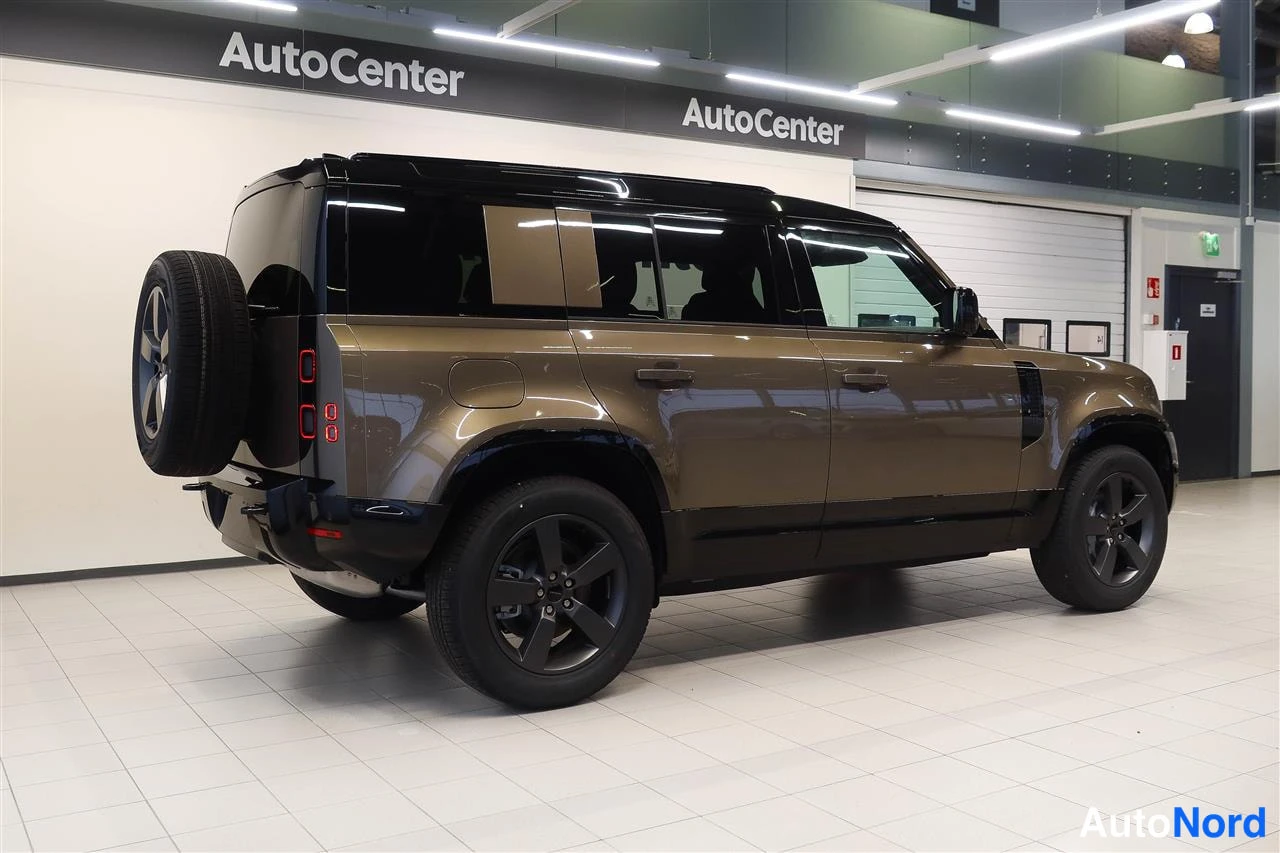 Takaviisto: Land Rover Defender -maasturin takakolmannes sisätiloissa, ruskeakarakkinen koriväri, takarenkaita kannattava varapyörä takaosassa ja mustat kevytmetallivanteet