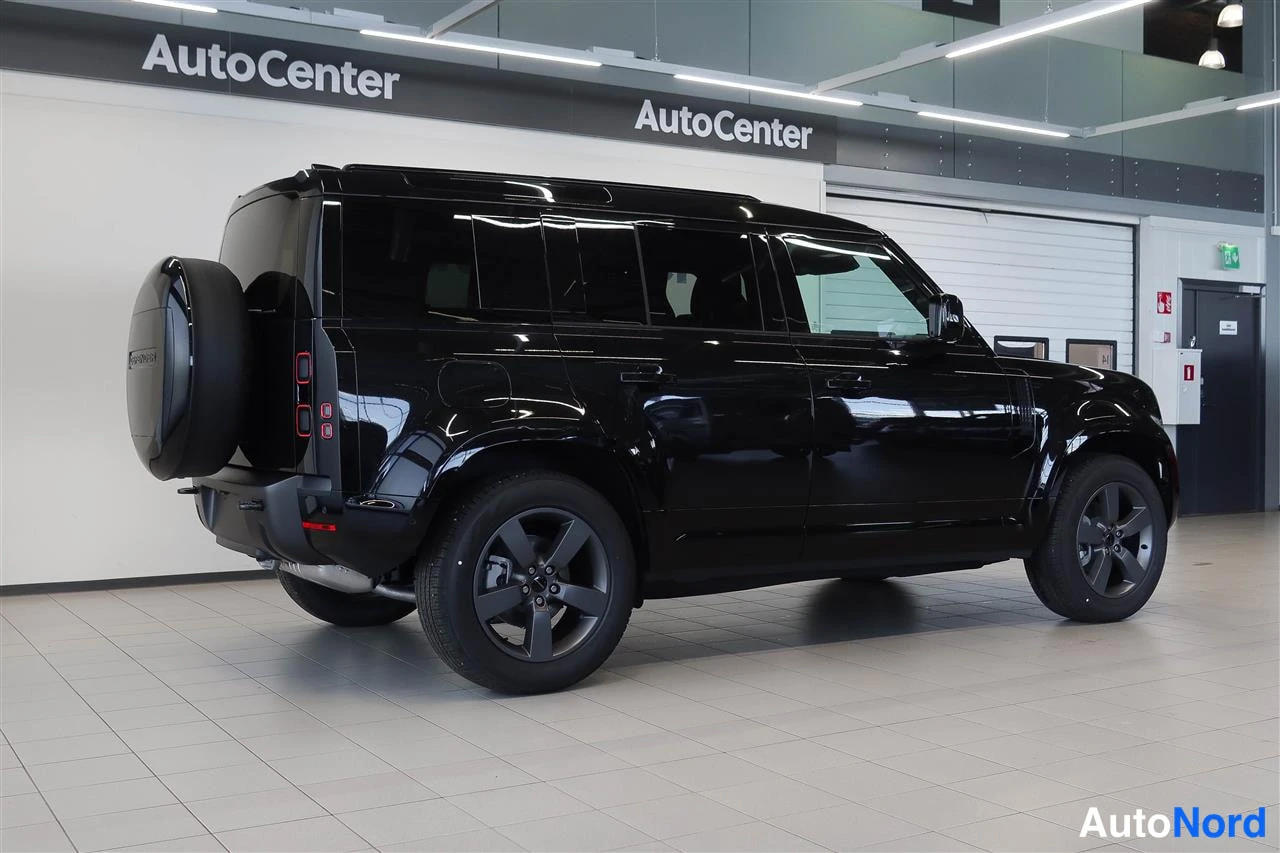 Takaviisto: Musta Land Rover Defender näkyvä takaa sivulta Autocenter-hallissa, varapyörä takaluukussa ja tummat vanteet.
