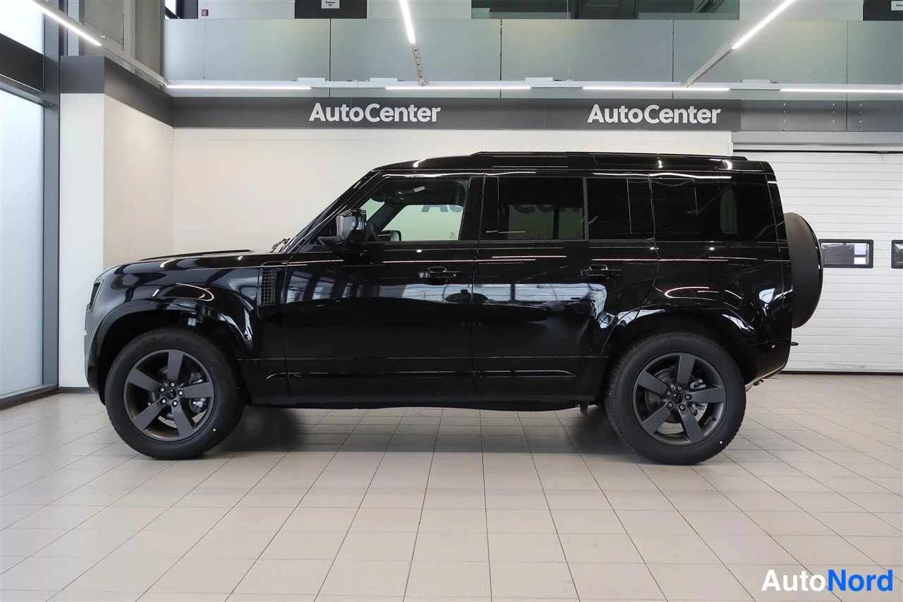 Sivulta: Musta Land Rover Defender sivulta kuvattuna Autocenter-hallissa, tummat alumiinivanteet ja varapyörä takana.