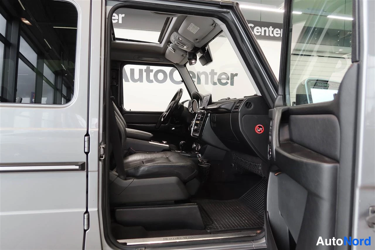 Muu: Kuljettajan oven auki: Mercedes-Benz G:n tumma kojelauta, ohjauspyörä ja istuimet kuvattuna auton kyljestä.