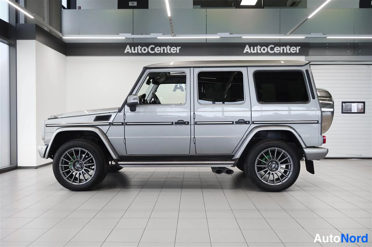 Sivulta: Hopeanharmaa Mercedes-Benz G sivulta kuvattuna autosalin sisällä; kyljen ovien ikkunat, kattorailot ja renkaat näkyvät.