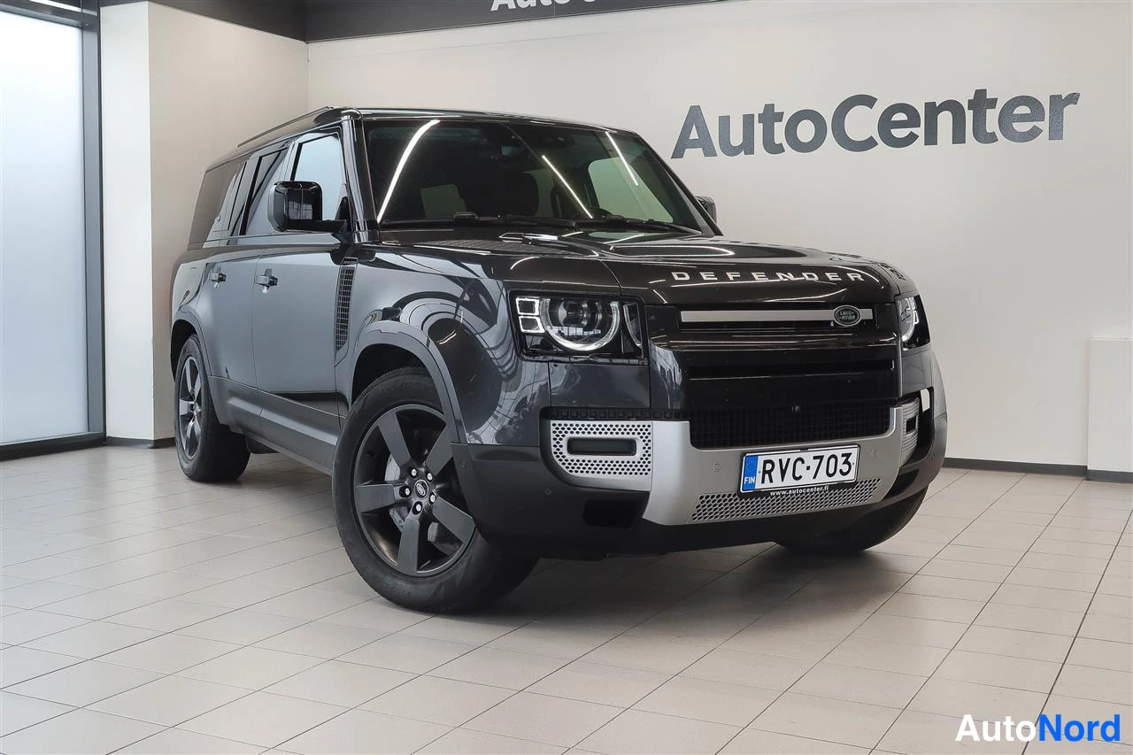 Etuviisto, nokka oikealle: Land Rover Defender, tumma, edessä oikealta vinasti kuvattuna autosalin lattialla.