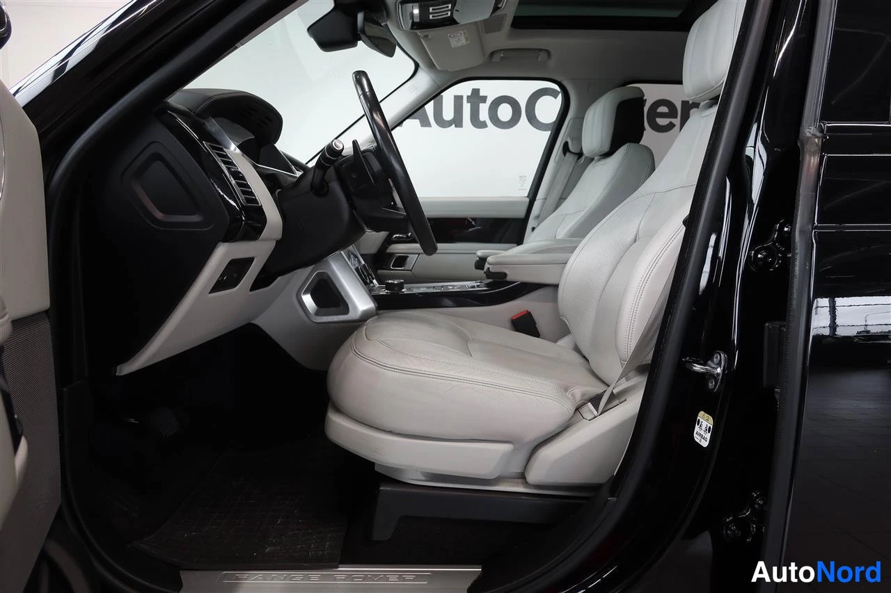 Muu: Avoin kuljettajan ovi sisätiloihin: beige nahkainen istuin, ohjauspyörä ja kojetaulu, taustalla AutoCenter-tunnus.