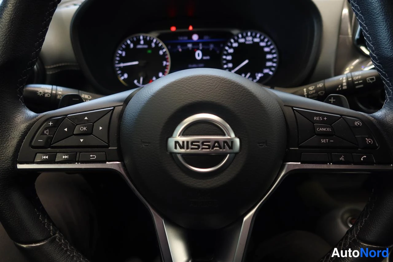 Takaviisto: Nissan-merkkinen ohjauspyörä lähikuvassa, puhelin- ja cruise-napit sekä kylkivaihteiston vivut näkyvät, mittaristo taustalla.