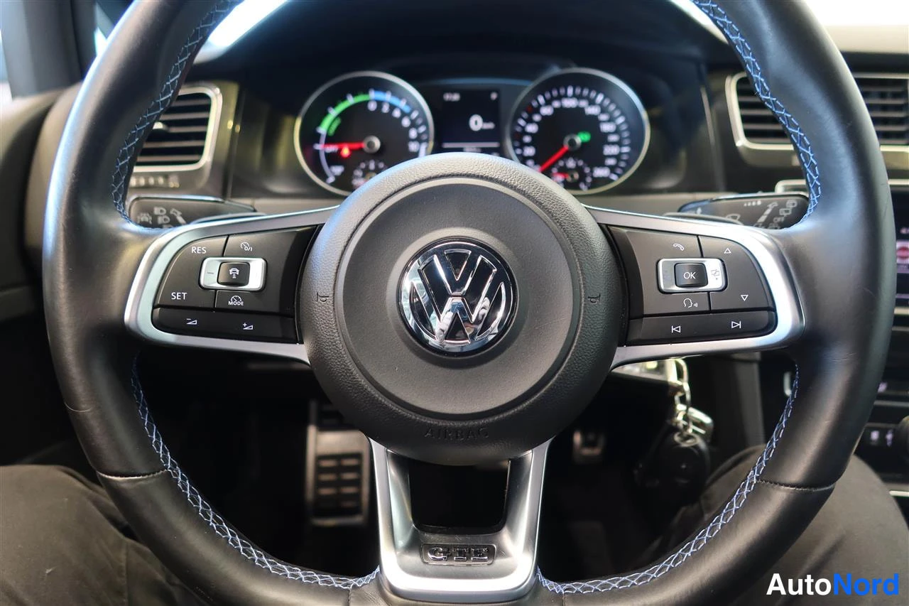 Muu: Lähikuva ohjauspyörästä, jossa VW-logo ja monitoiminapit, taustalla mittarit valaistuina.