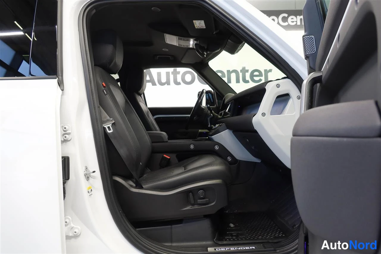 Yksityiskohta: Avattu kuljettajan ovi näyttää tumman sisustan: nahkapenkit, ohjauspyörä ja kojelauta, kynnyksellä 'DEFENDER'-teksti. Taustalla AutoCenter-mainos.