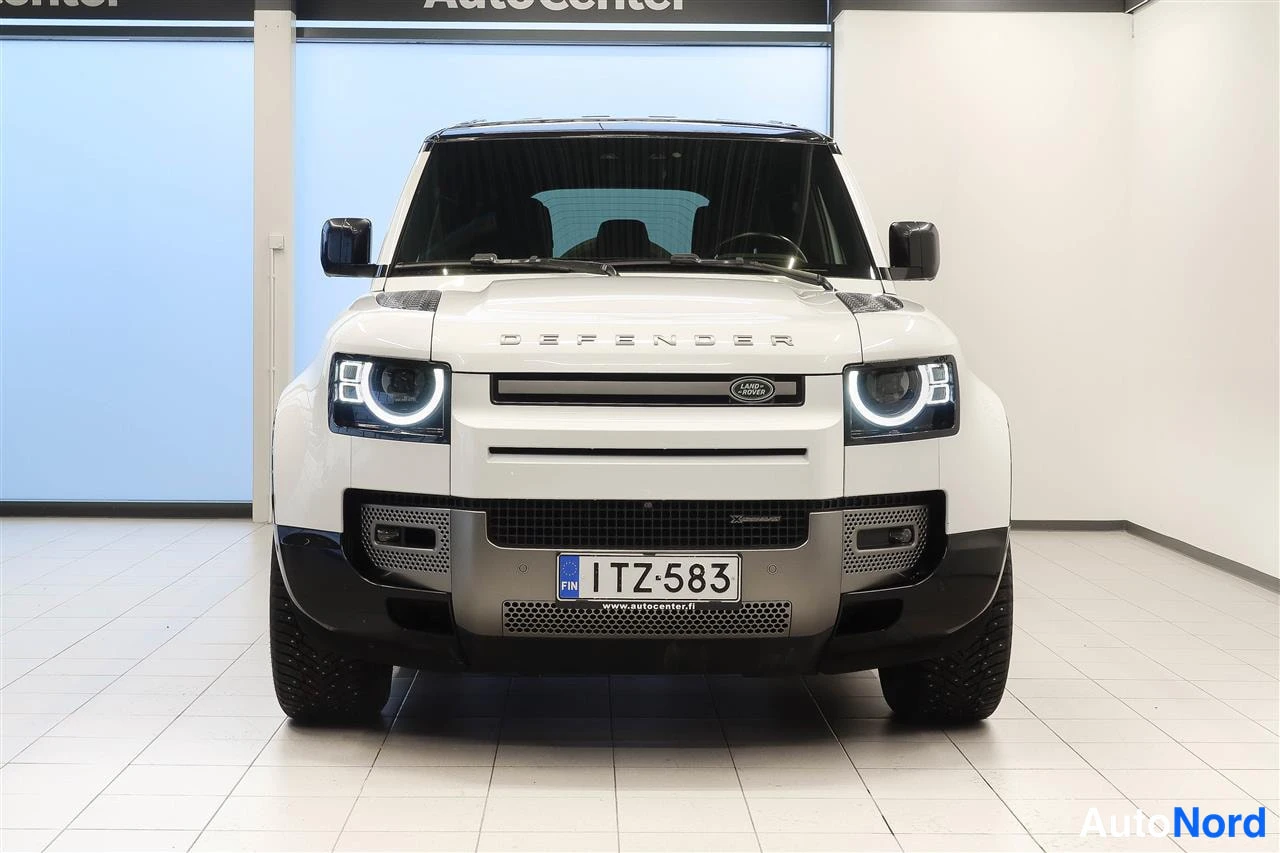 Edestä: Valkoisen Land Rover Defenderin etunäkymä suoraan edestä, LED-ajovalot päällä, rekisteritunnus.