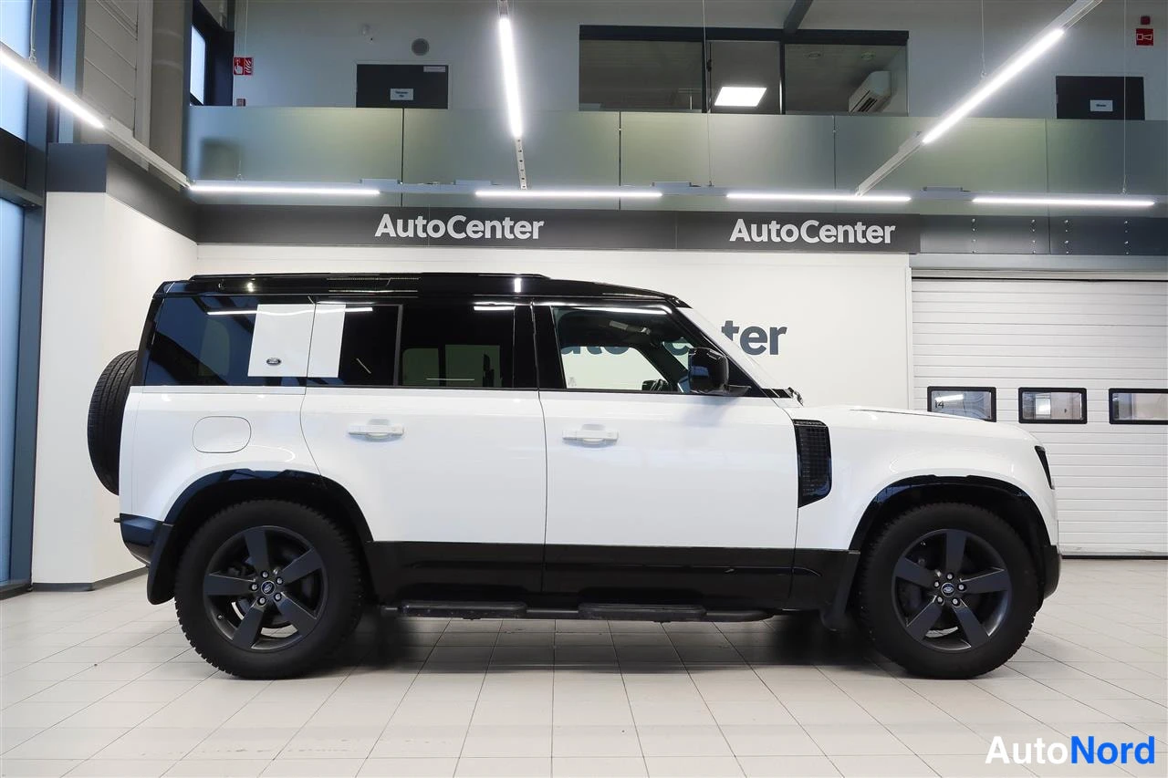 Sivulta: Valkoisen Land Rover Defenderin sivuprofiili vasemmalta, varapyörä takaluukulla, näyttelytilassa.