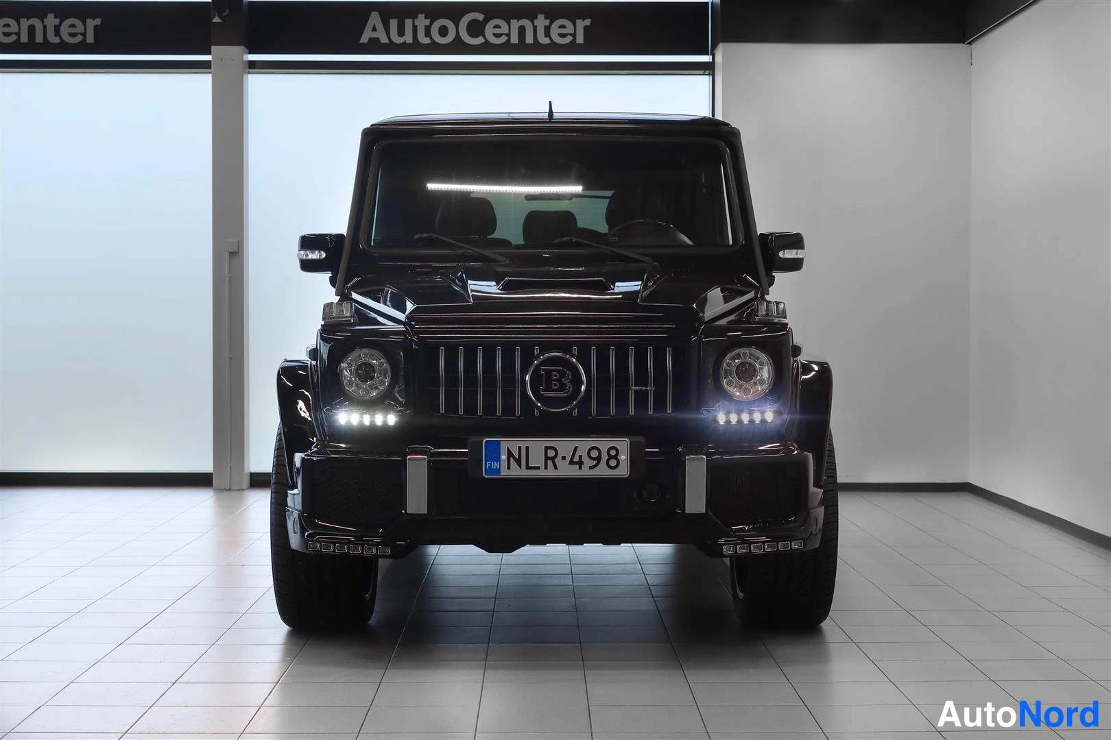 Edestä: Mustan Mercedes-Benz G:n etunäkymä suoraan edestäpäin, ajovalot päällä, rekisteri.