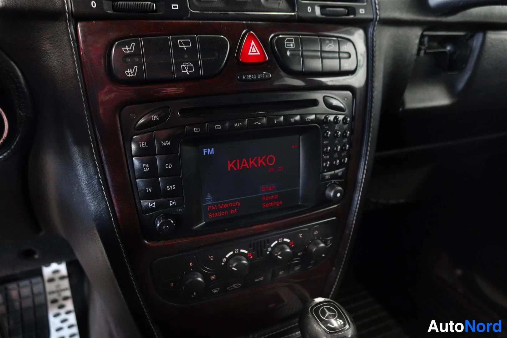 Yksityiskohta: Sisätilakuvan keskikonsoli: radio-navigointiyksikkö näytöllä 'KIAKKO' FM-tilassa, painikkeet TEL, NAV, CD, TV, SYS; ylhäällä varoitusvalot ja AIRBAG OFF -merkin
