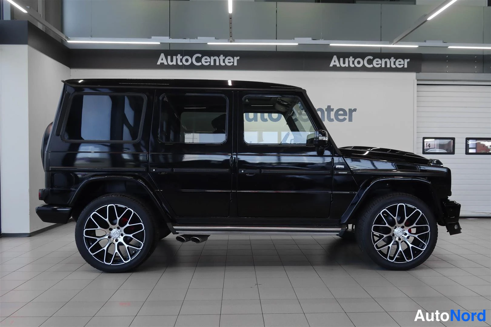 Sivulta: Musta Mercedes-Benz G -maasturi sivulta sisätiloissa, kolmiovinen takapää ja kattotelineet näkyvät.